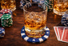 Glass of whiskey with ice on a poker-themed coaster surrounded by poker chips and cards.